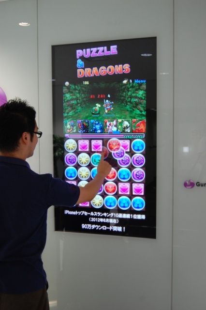 写真は2012年9月のインタビュー時の山本P。ガンホー本社受付にあるモニターでアーケード版パズドラごっこをしたのだが、ついに実現することになった！